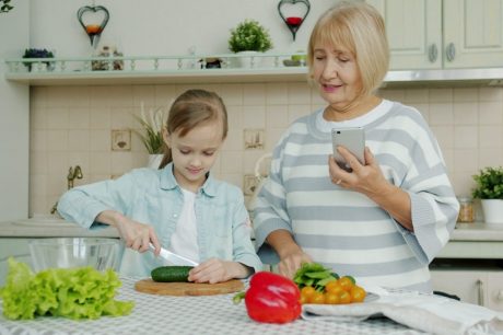 Grandmother with smartphone is teaching granddaughter to cook salad at home, cute little girl is cutting cucumber using knife and chopping board.